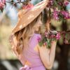 A woman in a purple dress and hat is looking at a tree with pink blossoms