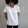 Young African American female in white t shirt with curly hair standing against gray wall and holding hands behind back and laughing happily with closed eyes